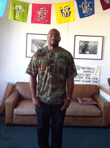 A picture of Taylor Code standing in front of a brown couch with a wall in the background that has two black and white framed pictures and turquoise, green, yellow, navy blue, and purple flags hanging from ceiling over his head.
