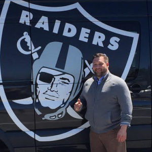 White man with beard and gray sweater staniding in front of Oakland Raiders logo.