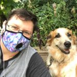 Image description: Skye, a young, disabled, white trans person, sits outside with one of their pups, Cricket, an orange and creme shepherd mix. Skye is wearing thin-rimmed rectangular eye glasses, small white headphones, a gray Henley shirt, and a blue, white, and pink swirly-styled (trans pride colours!) breathing mask. Cricket and Skye are both smiling and behind them is a mess of tall green plants.