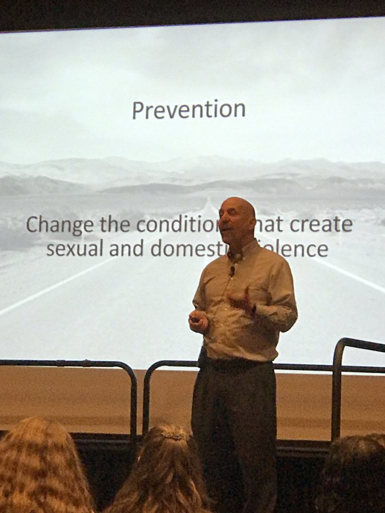 David Lee (balding white man in white shire and dark pants) standing in from on screen saying "Prevention: Change the conditions that create sexual and domestic violence"