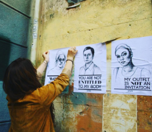 Women pasting up 3 "Stop Telling Women to Smile" posters. The center and the right poster have visible text. Center: black and white sketch of a women with short hair with the text "You are not entitled to my body." Right: black and white sketch of another woman with short hair with text "My outfit is not an invitation." For more information about International Anti-Street Harassment Week, go to http://www.meetusonthestreet.org/